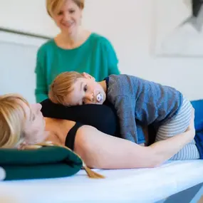 Physiotherapie: Mutter liegt auf Liege, Kind kuschelt, Therapeutin im Hintergrund